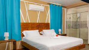 A hotel room with a wooden bed, white bedding, a wooden headboard, a nightstand with a lamp, a bathroom with a glass shower enclosure, and a towel on the bed. - The Habitat Sesel (Mahé Island)