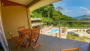 A balcony with a wooden table and chairs, a pool, and a view of lush greenery and a distant island. - The Habitat Sesel (Mahé Island)