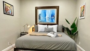 Et soveværelse med seng, natbord, lampe, en stor plante og billedkunst på væggene.