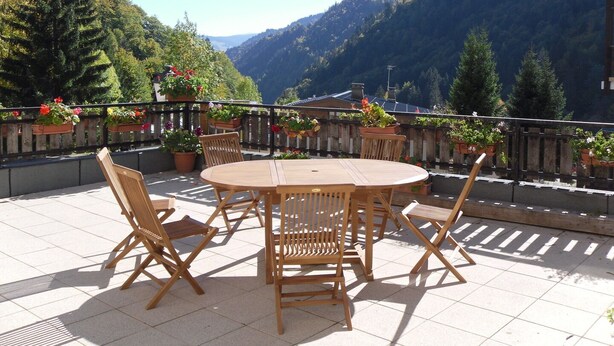 Eine Terrasse mit einem runden Holztisch und Stühlen, umgeben von Kübelpflanzen und einem Holzzaun.