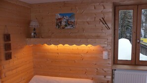 Ein Zimmer in einer Holzhütte mit einem Bett, einer Lampe, einem Bild von Hirschen an der Wand und Blick auf eine verschneite Landschaft durch eine Tür.