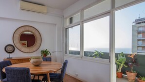 Uma sala de jantar com uma mesa redonda de madeira, cadeiras estofadas azuis, um espelho trançado e plantas em vasos.