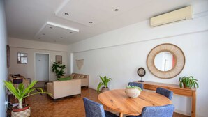 Uma sala de estar moderna com uma mesa de jantar de madeira, cadeiras azuis e um sofá bege. Há plantas em vasos e um espelho grande nas paredes.