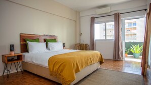 Quarto com cabeceira de madeira, cama com travesseiros brancos e verdes, manta cor mostarda, mesa de cabeceira com um alto-falante e janela com cortinas.