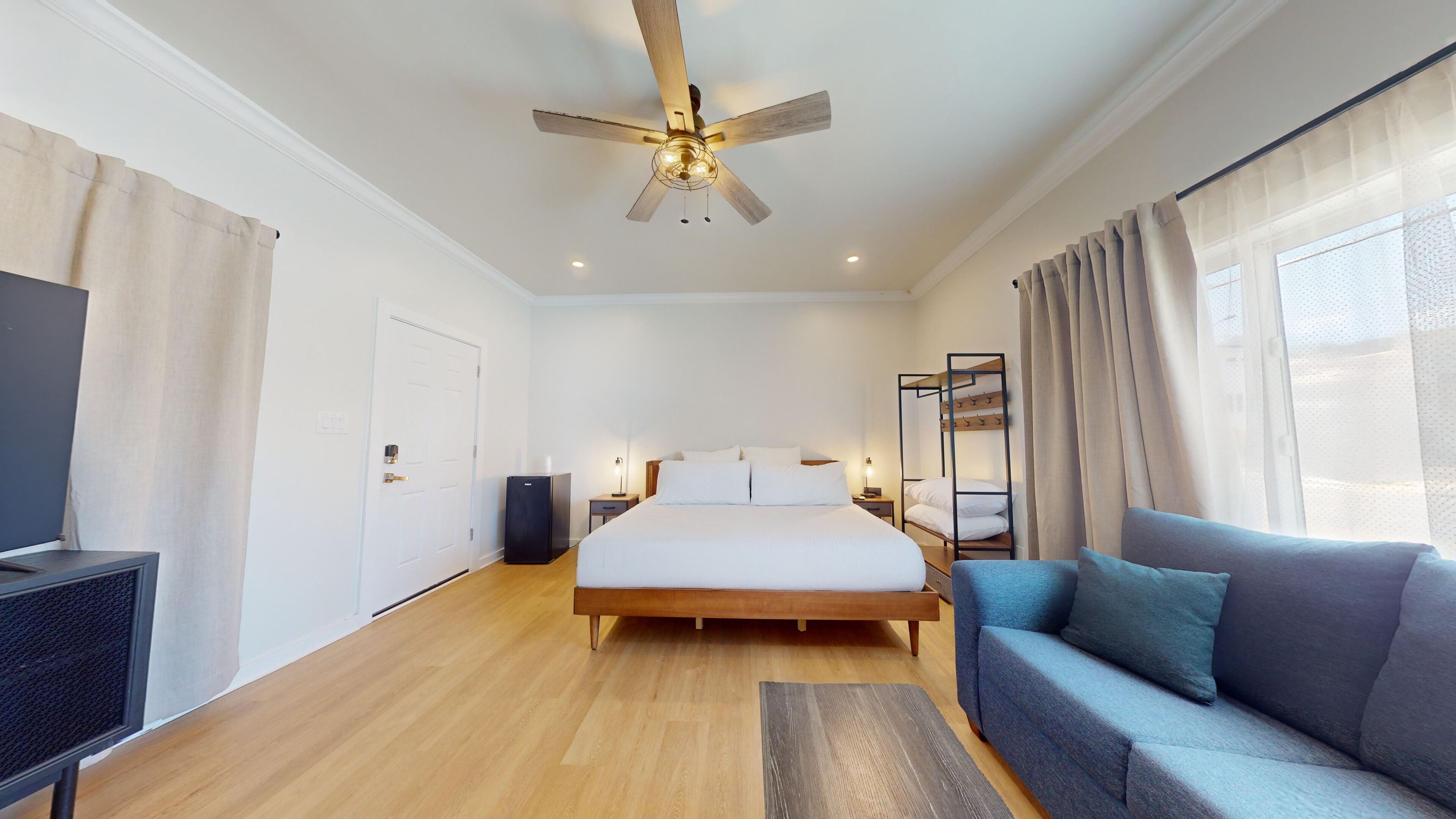 A modern bedroom with a wooden floor, a bed with white bedding, a ceiling fan, a TV, a sofa, and a window with curtains.