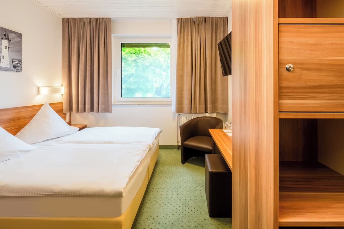 A hotel room with a bed, a desk, a chair, a wardrobe, a window with curtains, a lamp, and a wall-mounted TV.