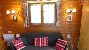 Una habitación acogedora con un sofá gris, cojines rojos estampados, paredes de madera y dos lámparas fijadas a la pared.