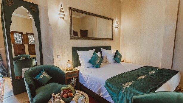 Una habitación de hotel con una cama grande, un sillón verde, una mesita de noche con una lámpara y un espejo en la pared.