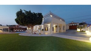 A modern house with a white facade, a large tree in the front yard, and a swimming pool in the background.