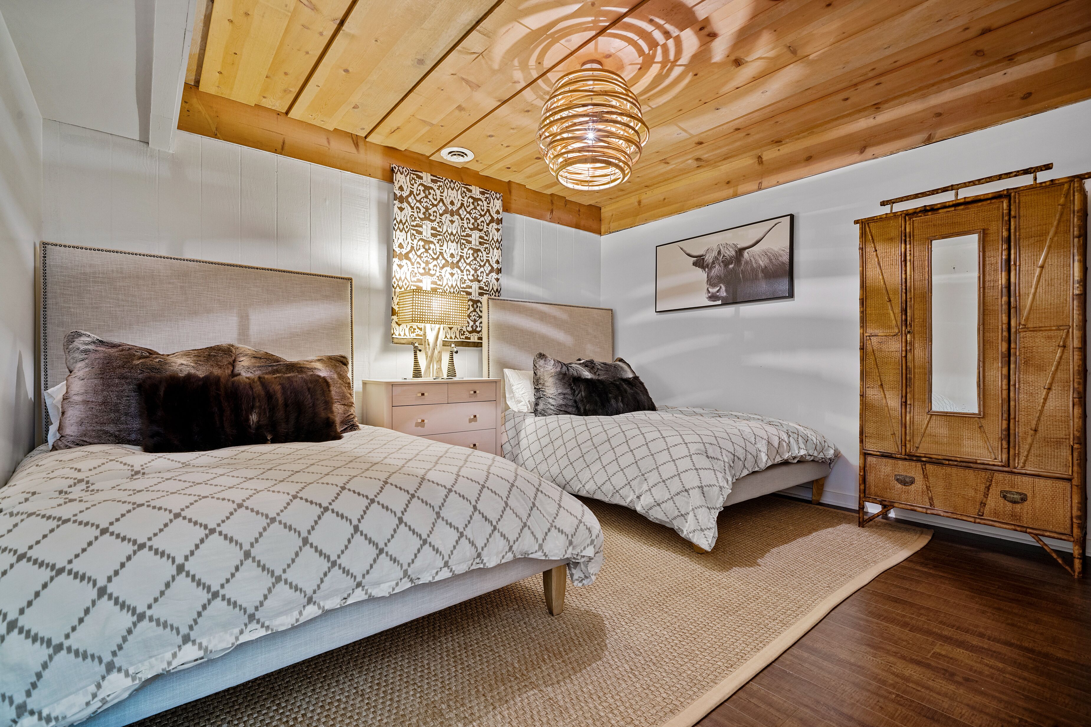 A bedroom with two beds, a wooden ceiling, a woven rug, a dresser, and a wall-mounted lamp.