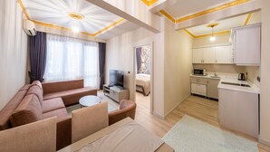 Un salon moderne comprenant un canapé, un téléviseur et un coin repas. Il y a une cuisine équipée de placards et d’un four à micro-ondes. Le plafond de la pièce est décoré de motifs et un luminaire y est installé.