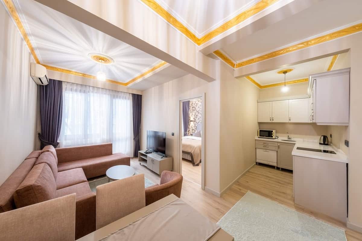 A modern living room with a sofa, TV, and dining area. There is a kitchen with cabinets and a microwave. The room has a ceiling with decorative patterns and a light fixture.
