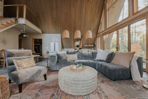 A spacious living room with a large gray sectional sofa, a round ottoman, and a high ceiling with wooden beams. - *NEW* Luxury A-Frame by Sunday River w/ hot tub (Bethel)