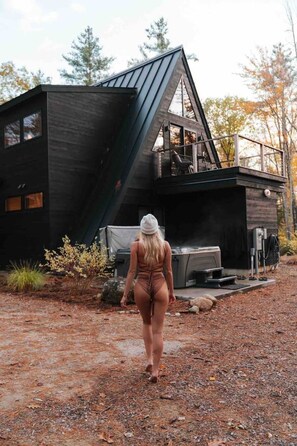 A person walking towards a modern, dark-colored cabin with a sloped roof, a deck, and a covered outdoor area. - *NEW* Luxury A-Frame by Sunday River w/ hot tub (Bethel)