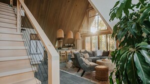 A spacious living area with a high ceiling and wooden beams, a modern kitchen, and a large window allowing natural light. - *NEW* Luxury A-Frame by Sunday River w/ hot tub (Bethel)