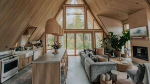 A spacious loft with a high ceiling, wooden beams, a kitchen with modern appliances, a dining area, a living room with a sofa and large windows offering a view of the outdoors. - *NEW* Luxury A-Frame by Sunday River w/ hot tub (Bethel)