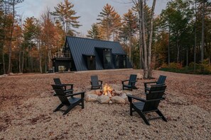 A cabin with a sloped roof surrounded by a gravel area with four Adirondack chairs arranged around a central fire pit. - *NEW* Luxury A-Frame by Sunday River w/ hot tub (Bethel)