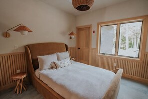 Une chambre avec un grand lit, deux lampes fixées au mur, une fenêtre donnant sur des arbres et une porte.