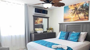 Un dormitorio con cama, una silla, ventilador de techo, televisión y obras de arte en la pared.