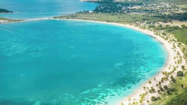 Vista aérea de una zona costera con playa, aguas cristalinas y un pequeño asentamiento al fondo.