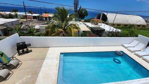 Una piscina en la azotea con sillones de descanso, una parrilla y vistas al océano y edificios.