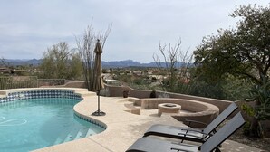 A swimming pool with a tiled edge, lounge chairs, a fire pit, and a view of mountains and houses in the background.
