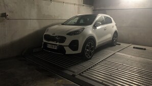 Um SUV Kia branco estacionado em uma garagem subterrânea com pouca luz, paredes de concreto e uma rampa de metal.