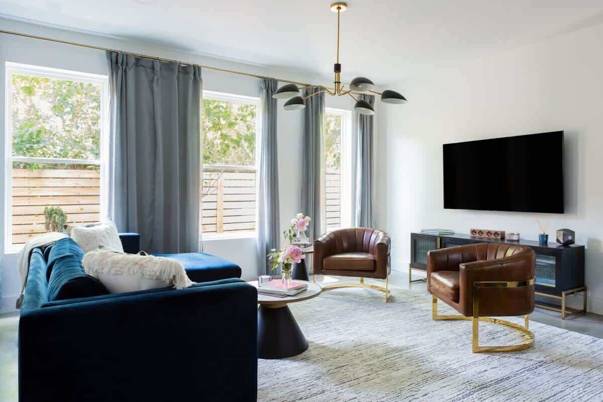 Een moderne woonkamer met een blauwe bank, een bruine leren fauteuil en een salontafel. Een flatscreen-tv hangt aan de muur, en er is een groot raam met gordijnen.