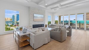 Amplia sala de estar con televisor grande, asientos cómodos, pared de acento de piedra y ventanas amplias con vista a la playa y edificios.