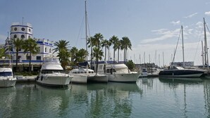 A marina with several yachts docked, a building with a tower, and palm trees in the background. - 2-Bedroom Apartment with Direct Beach Access & Private Parking – Estepona (Estepona)