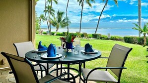 Een ronde tafel gedekt voor twee personen, met stoelen, een groen zonnescherm en uitzicht op de oceaan en palmbomen.