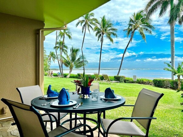 A round table set for two with chairs, a green awning, and a view of the ocean and palm trees.