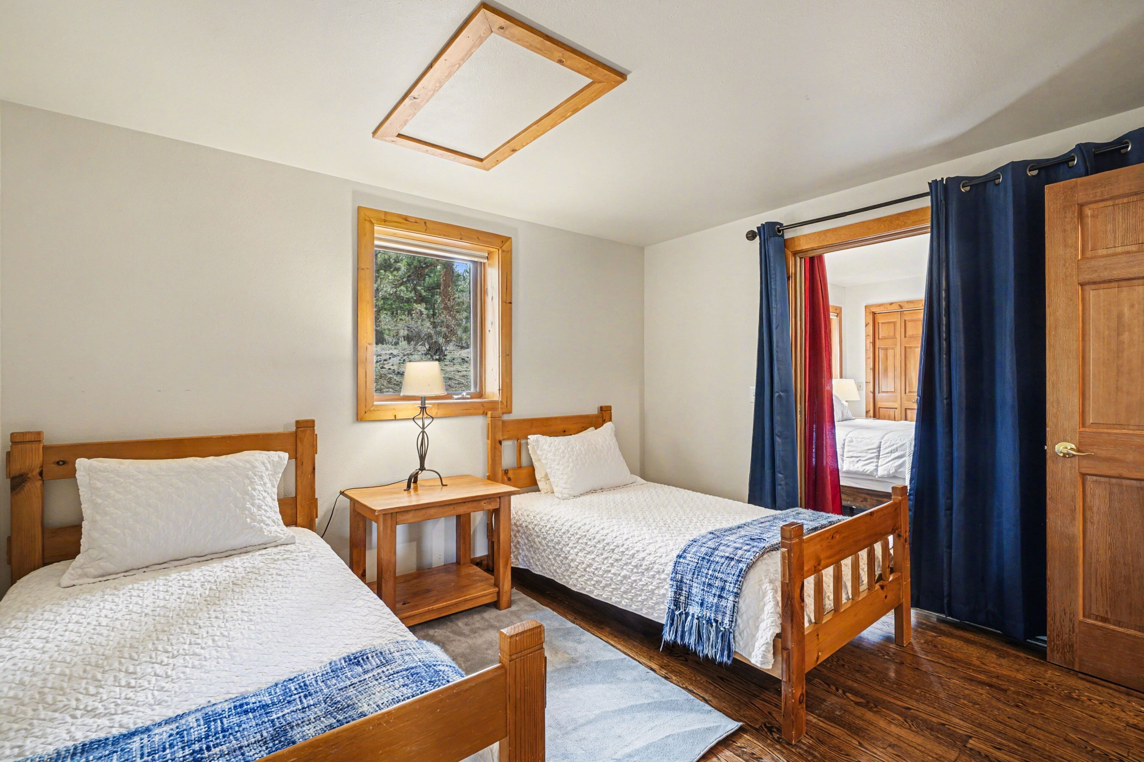Une chambre avec deux lits en bois, une petite table avec une lampe, une fenêtre donnant sur des arbres et une porte avec des rideaux bleus.