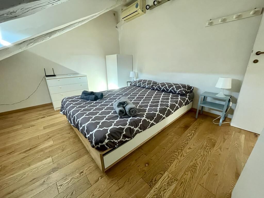 A bedroom with a bed, wooden flooring, a desk, and a lamp.