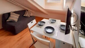 A compact room with a dining table set for two, a black sofa with striped cushions, a small window, and a television on a stand. - Casa Breton (Turin)
