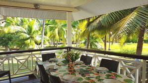Un área de comedor al aire libre cubierta, con una mesa dispuesta para cuatro personas, rodeada de sillas y vistas a un jardín tropical con palmeras.