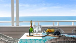 Una mesa con una botella de champán, copas y un frutero en un balcón con vista al océano.