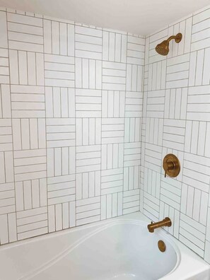 Un baño con un patrón de baldosas blancas tipo espiga, una bañera blanca y un cabezal de ducha dorado.