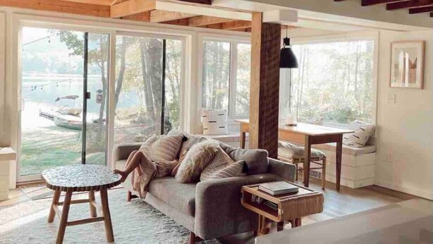 A cozy living room with a sofa, a small round table, and a view of a lake through large sliding glass doors. - Maine lake front ski getaway! (Greenwood)