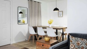 Uma sala de estar moderna com área de jantar, paredes brancas, um espelho, uma luminária pendente preta e um sofá com almofada estampada.