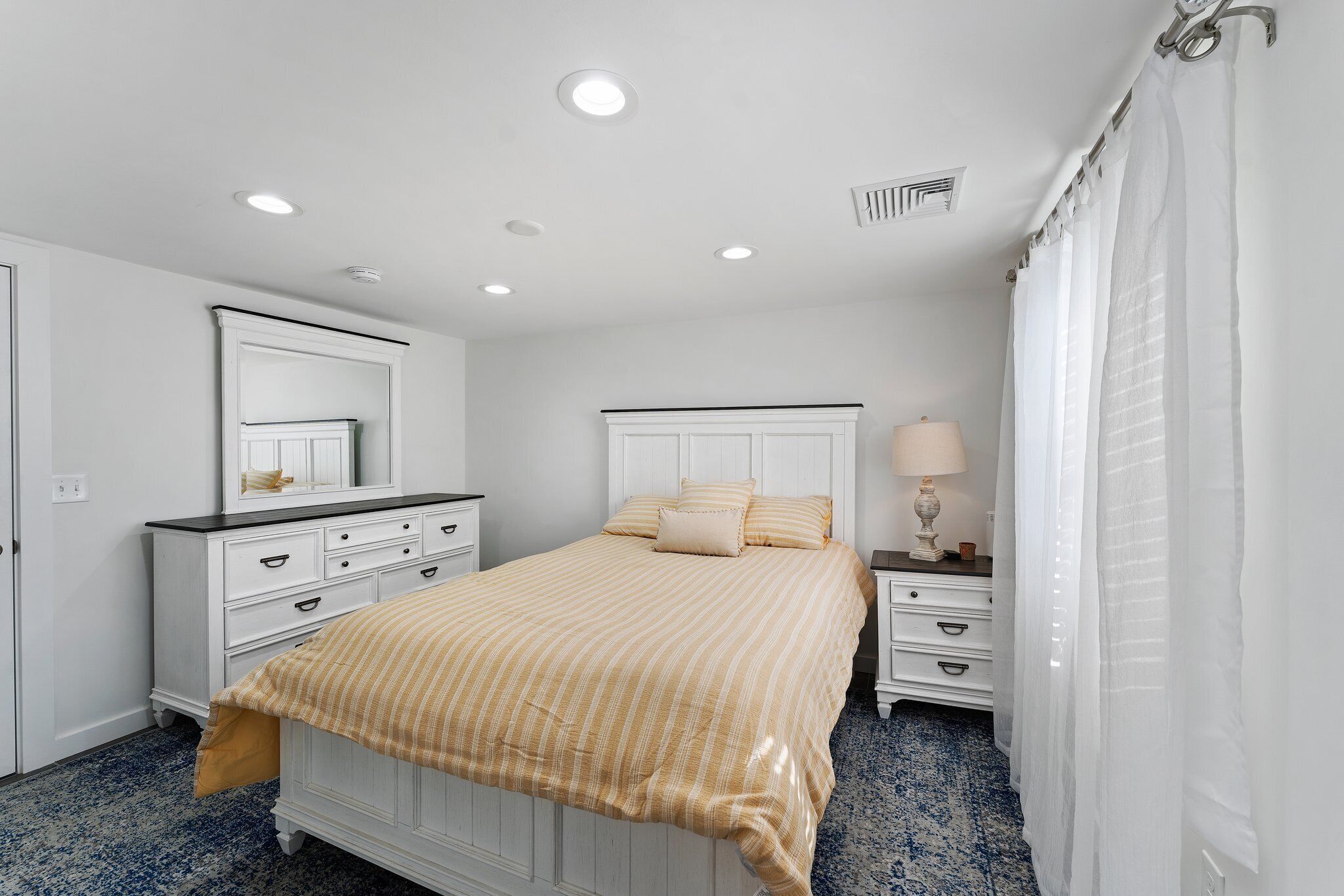 A bedroom with a bed, a dresser, a nightstand, a mirror, and a window with curtains.