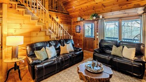 Ein gemütliches Blockhauss-Interieur mit einer Holztreppe, zwei Ledersofas mit Kissen, einem kleinen runden Tisch mit Tablett und einer Lampe.