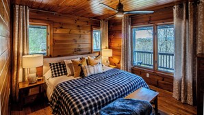 Ein gemütliches Schlafzimmer im Blockhausstil mit einem großen Bett, zwei Nachttischlampen, einem hölzernen Kopfteil und Blick ins Freie durch zwei Fenster.