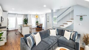 A modern living room with a grey sectional sofa, a kitchen with white cabinetry, and a staircase with white railings. - Hot Tub • Porches • Parking • Walk to King St! (Charleston)
