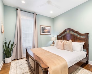 Un dormitorio con una cama de madera, una mesita de noche con lámpara, una ventana con cortinas translúcidas y un cuadro en la pared.