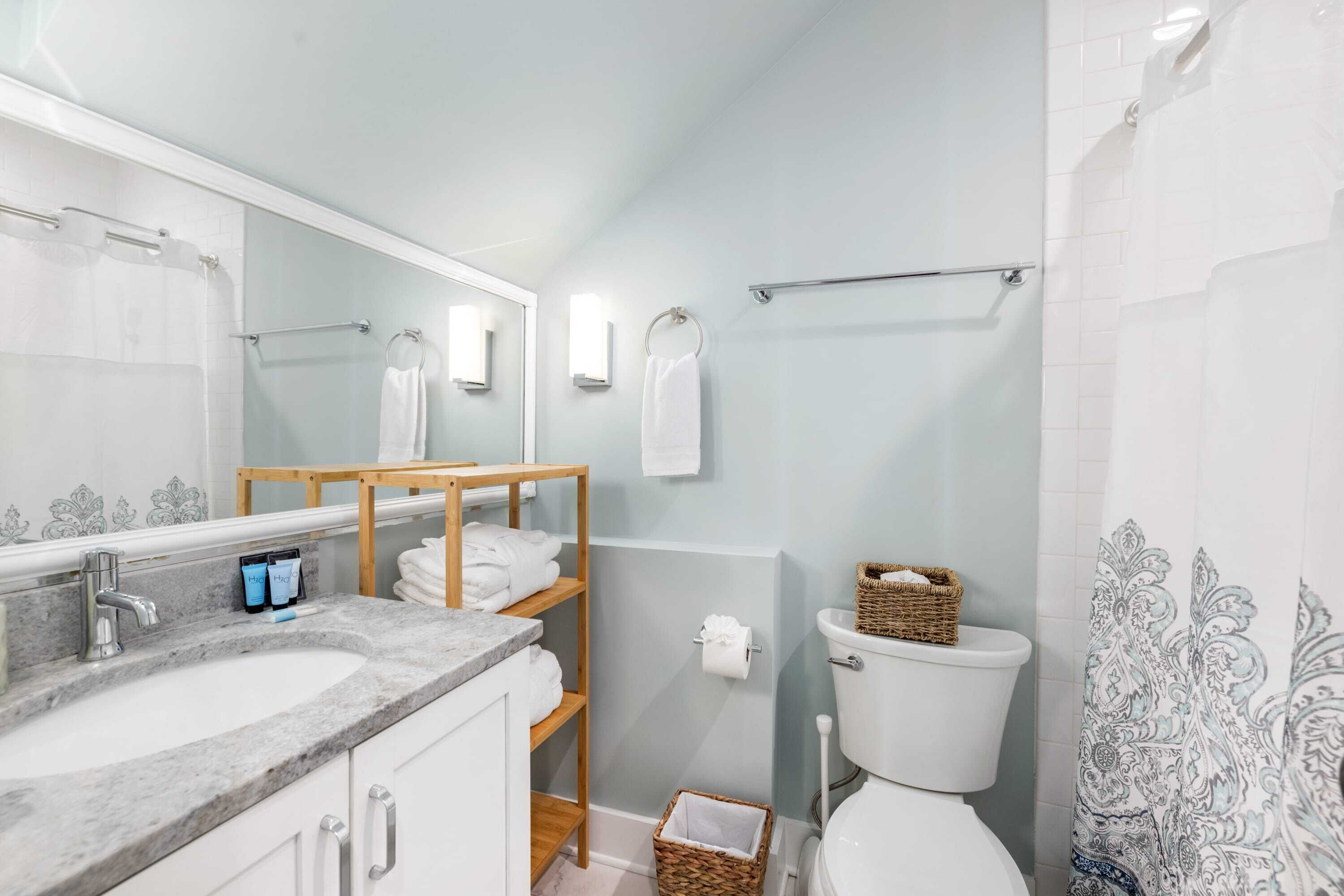 A bathroom with a marble sink, a wooden shelf with towels, a toilet with a basket, and a patterned shower curtain.