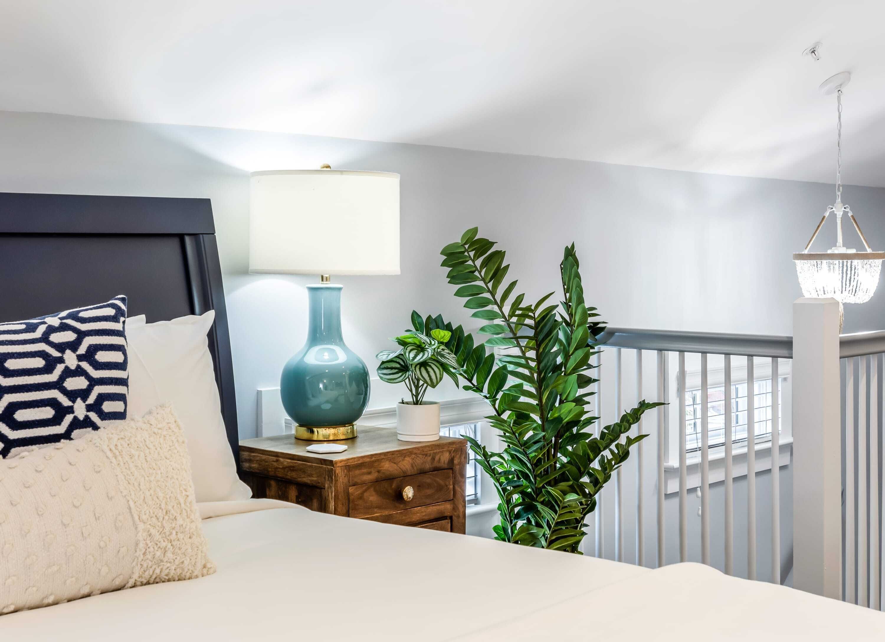 A bedroom with a bed, a nightstand, a lamp, a potted plant, and a view of the outside.