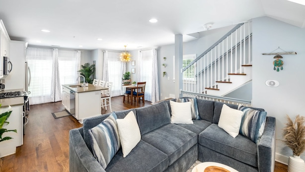 A modern living room with a grey sectional sofa, a kitchen with white cabinetry, and a staircase with white railings. - Hot Tub Escape • Parking • Walk to King St! (Charleston)