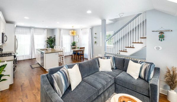 A modern living room with a grey sectional sofa, a kitchen with white cabinetry, and a staircase with white railings.