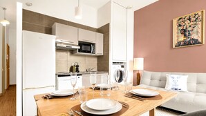Una cocina moderna con electrodomésticos blancos, un comedor con mesa de madera dispuesta para cuatro personas, y una sala de estar con sofá y un cuadro en la pared.